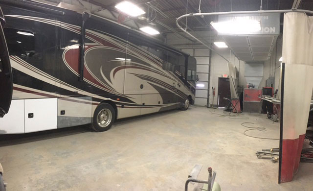 The image shows a large motorhome parked inside a building with an industrial setting, featuring metal structures and equipment in the background.