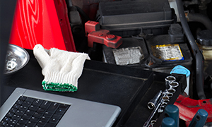 An open car hood with various tools and parts visible, including gloves on the laptop keyboard.