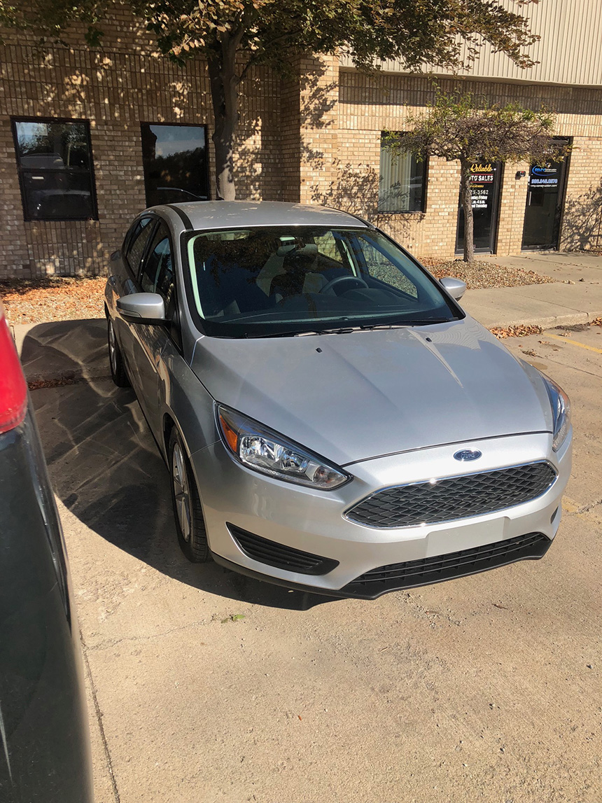 The image shows a silver Ford car parked outside a building with a person visible inside the vehicle.