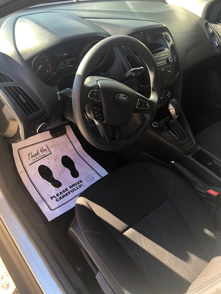 The image depicts the interior of a car with a focus on the driver s seat area, featuring a steering wheel, gear shift, and a sign with footprints placed on the front passenger seat.