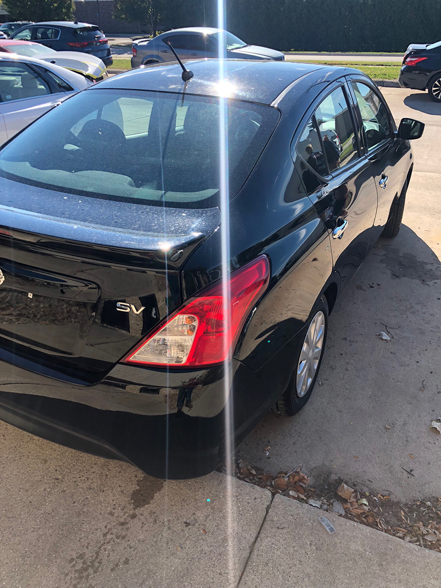 The image shows a black sedan parked outdoors during daylight, with a reflection visible on its surface.