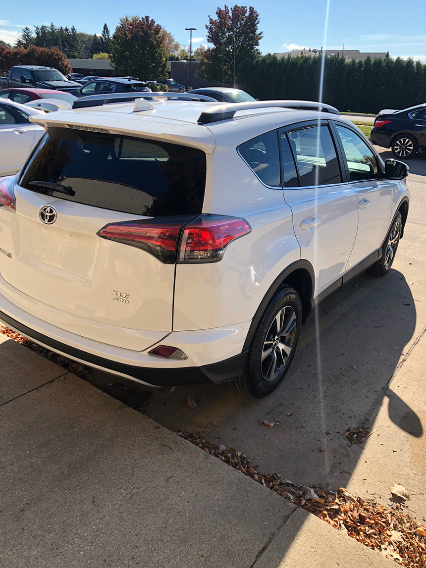 The image shows a white Toyota SUV parked at an angle next to a paved surface with trees in the background, under a clear sky.